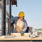 A female carpenter uses a power drill at an outdoor construction site.