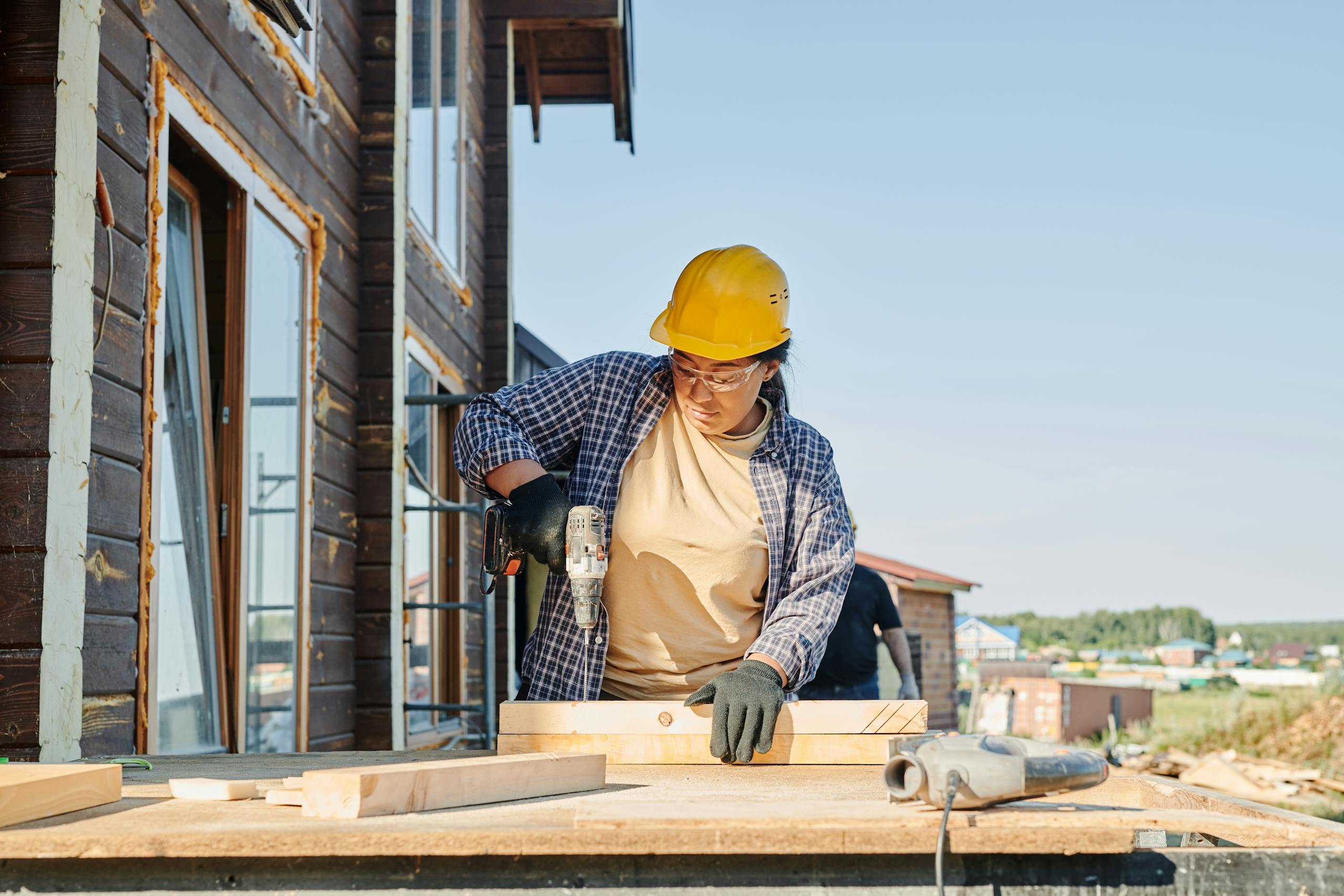 A female carpenter uses a power drill at an outdoor construction site.