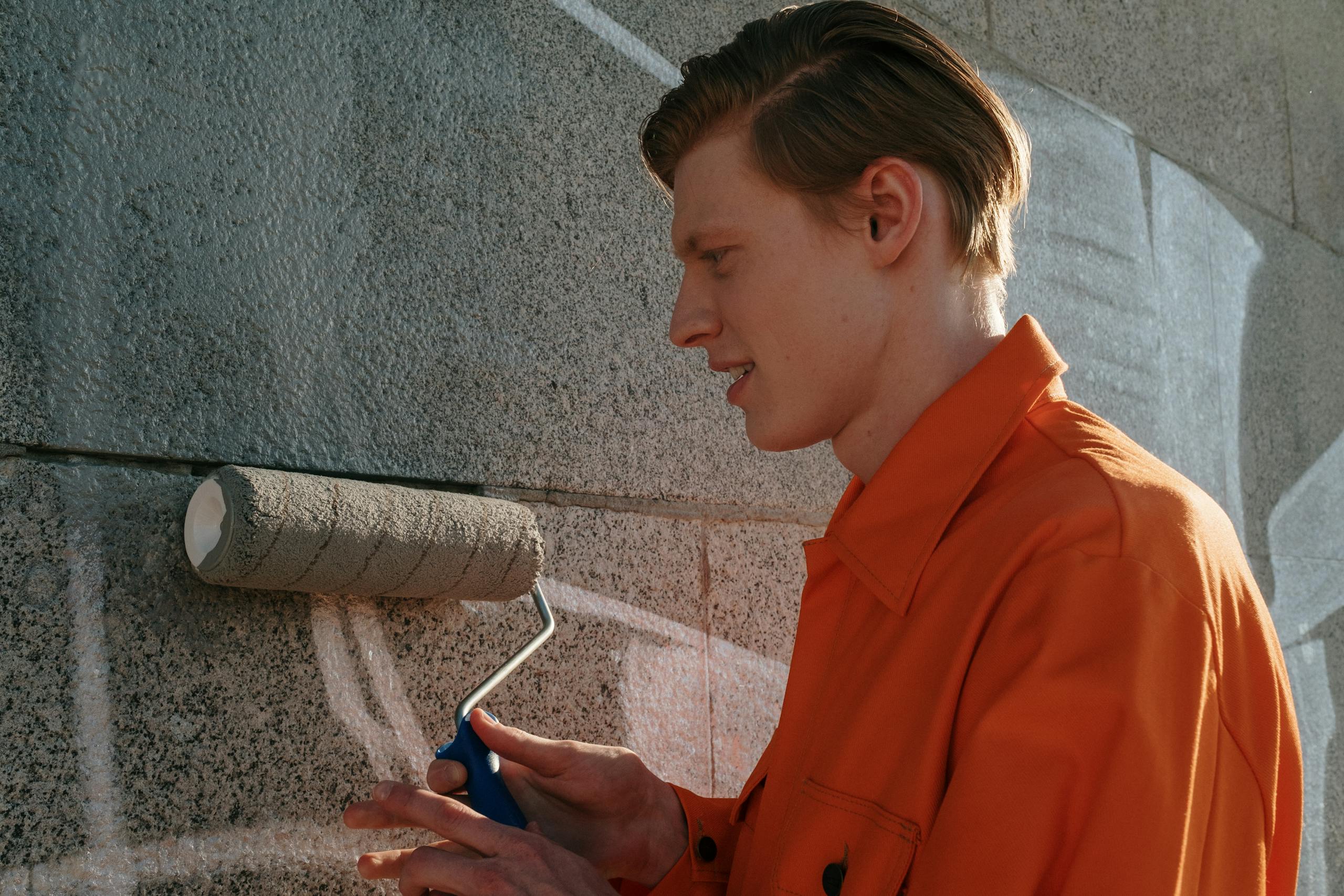 Young adult participating in community service, painting over graffiti on a concrete wall.